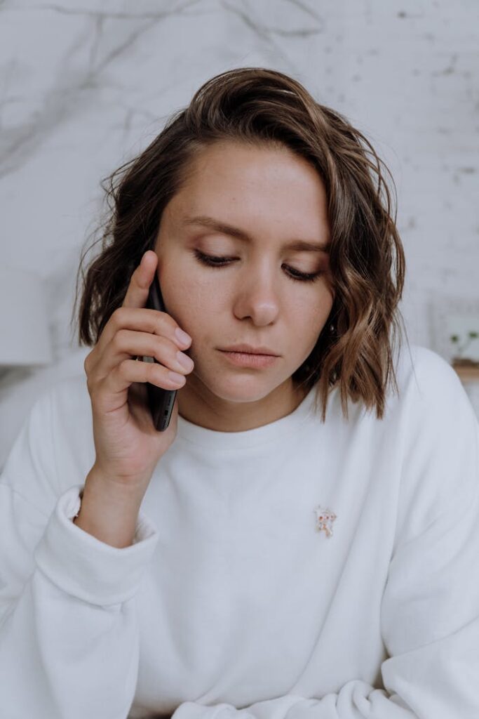 Young woman with bob haircut in white sweater talks on cellphone indoors.